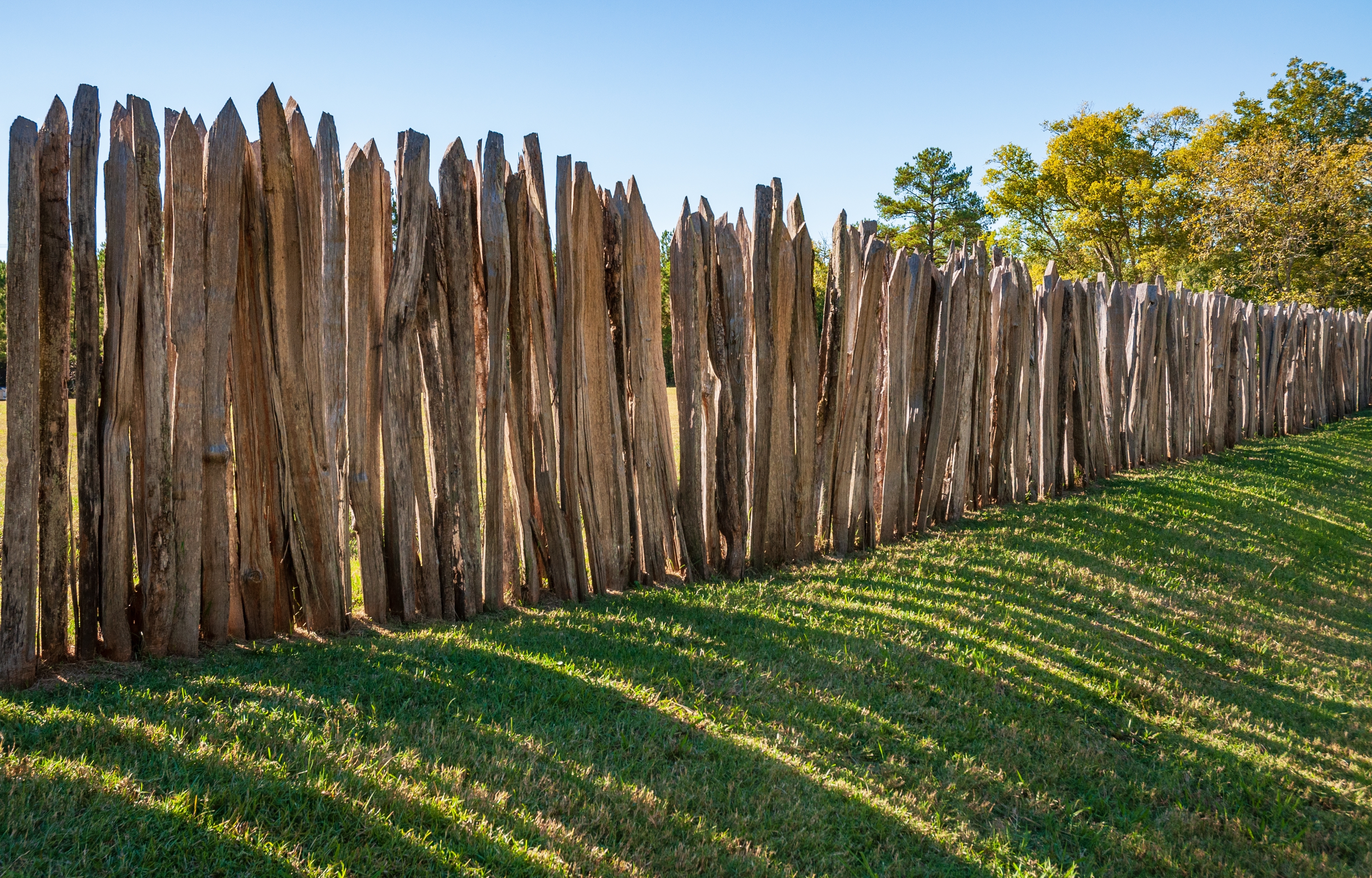 NinetySixNationalHistoricSiteOldNinetySixAndStar LKN Law Reconstructed tall pointed log palisade enclosing a grassy area with a wooden gate frame on an earthen berm framed by green and yellow trees against blue sky at Ninety Six